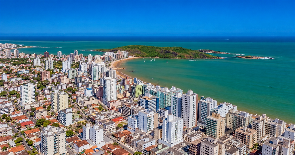 Vista aérea da Praia do Morro, em Guarapari, com prédios da orla, a enseada de água esverdeada e o morro coberto de vegetação ao fundo